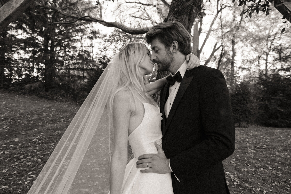 A bride and groom embracing and kissing under trees in Iowa, both in wedding attire, captured in a black and white photograph.