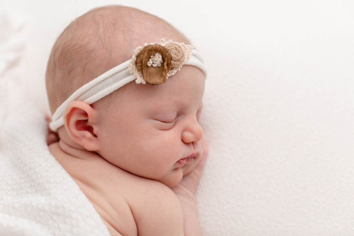 Baby girl sleeping on her belly with hand up by face. It is a close-up image of her face. She is wearing a headband with some small neutral flowers on it. She is laying on a white blanket and has a textured blanket draped over her back.