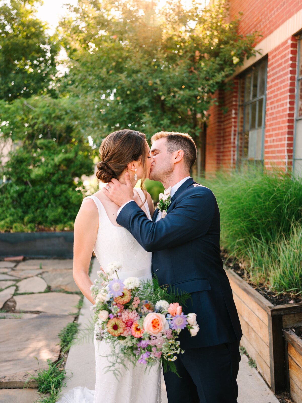 Bride and groom kiss outside of their wedding venue with her colorful Spring bouquet.