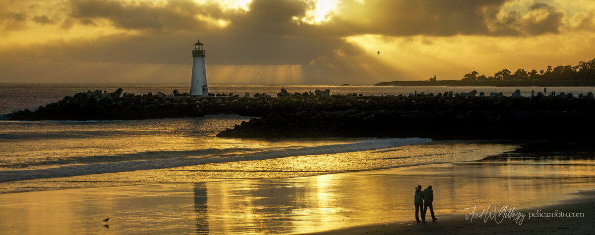 Couple walking along beach at sunset with Santa Cruz Harbor’s Walton Lighthouse and breakwater in horizon.