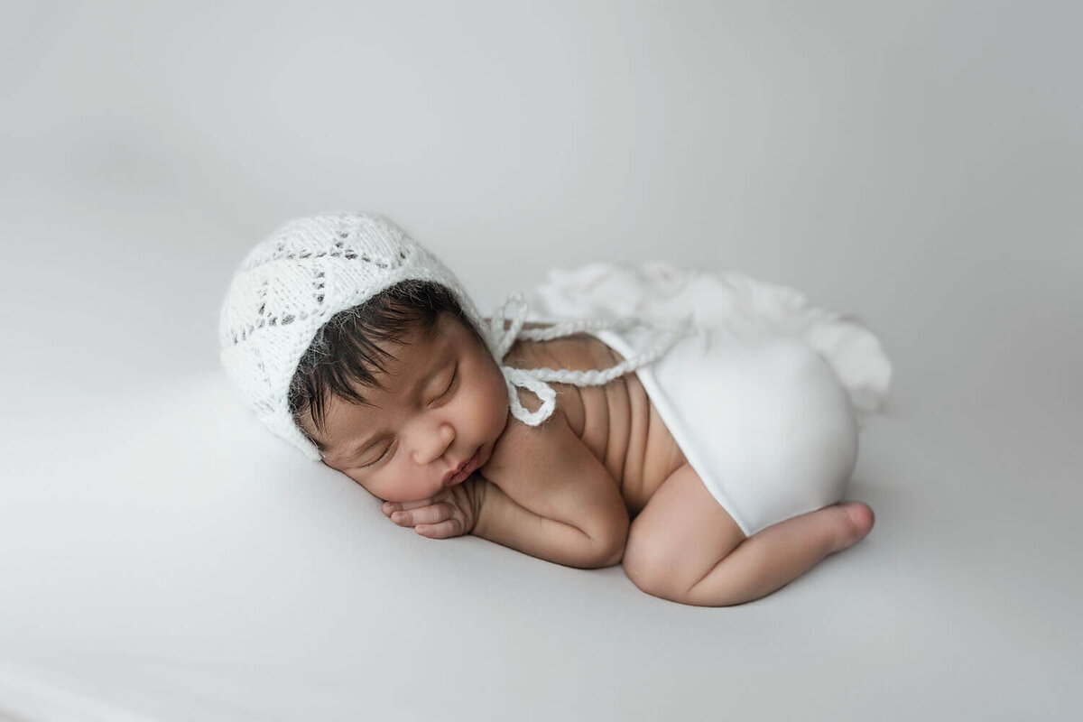 A newborn baby in a white knit bonnet sleeps on a white pad