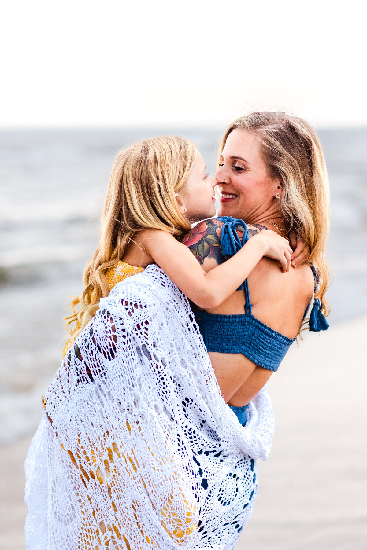 A beautiful, tender moment between mother and daughter wrapped in a soft lace blanket.  Their faces glow with love as they share a quiet hug by the ocean during a Virginia Beach family session with Brooke Tucker photography.