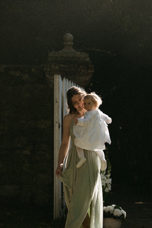 Une jeune femme en robe verte porte un bébé vêtu de blanc en souriant sous un rayon de soleil. Mariage au Château de Beaussais en Bretagne, capturé par Kamescop.