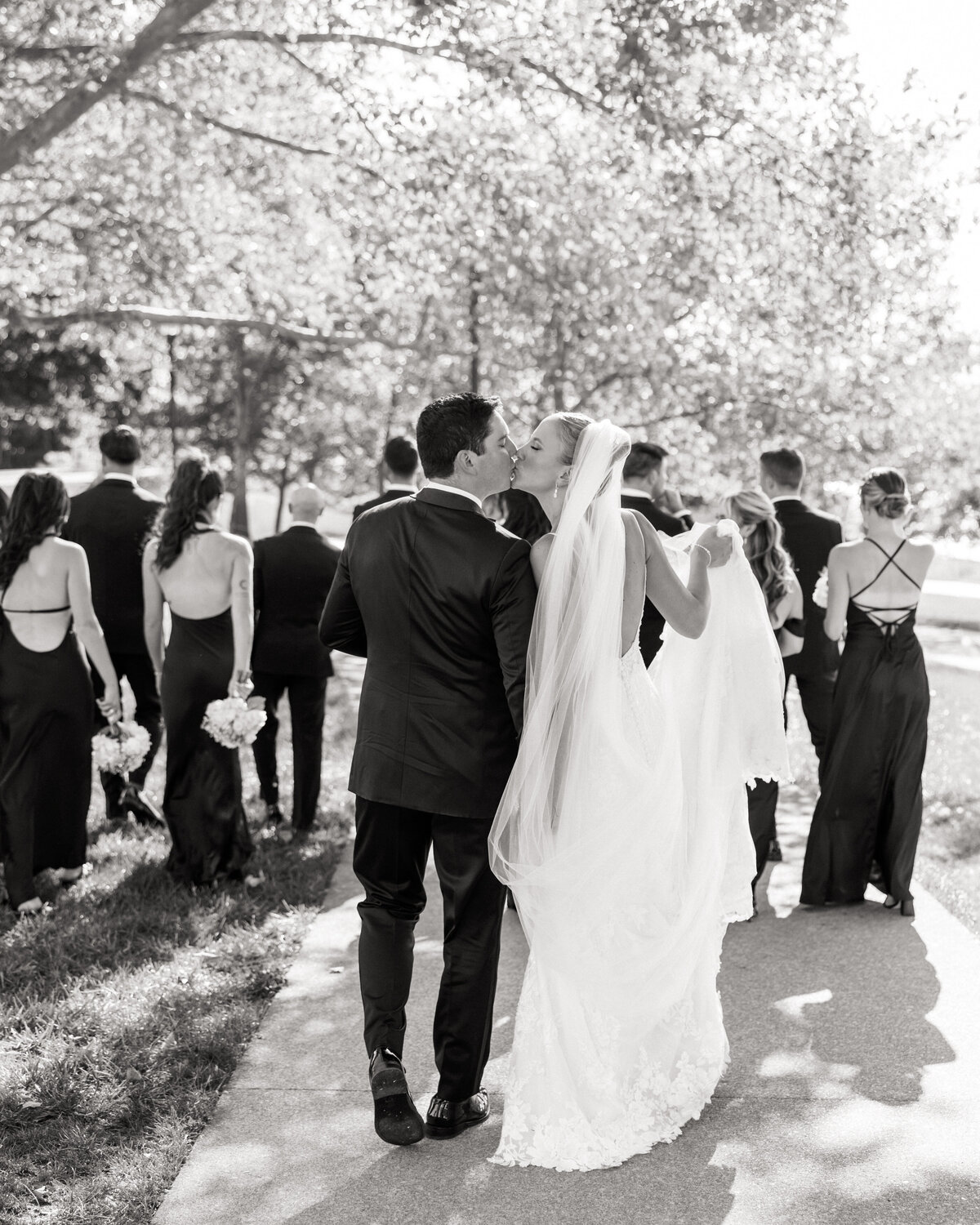 A romantic black and white moment as the bride kisses the groom while walking with their bridal party, snapped candidly by a Philadelphia wedding photographer.
