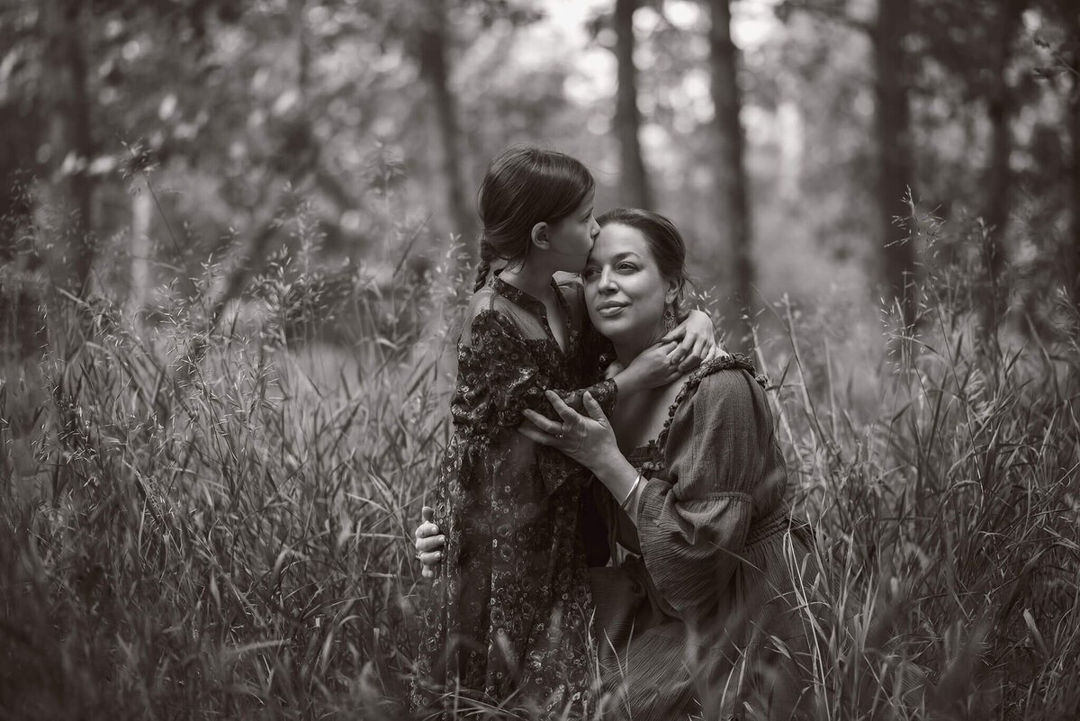 A black and white portrait of a mother and daughter embracing eaachother They are crouched down in tall grass. Taken by Brittany Danielle Photography