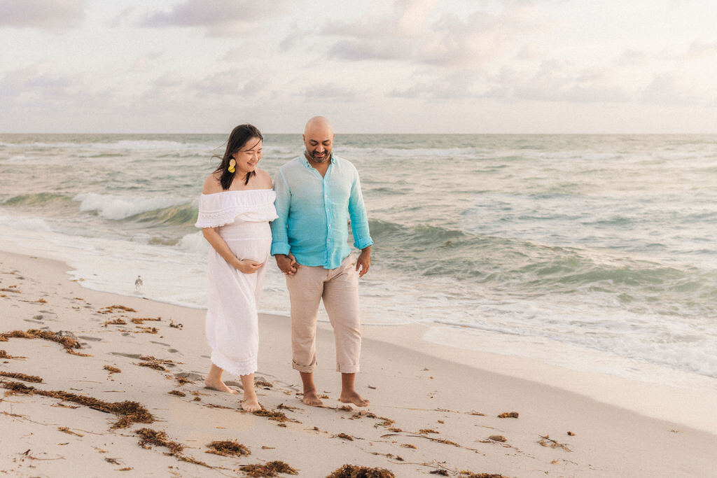 Pregnant couple walking barefoot along the shoreline during a maternity session at Boca Raton beach, South Florida.