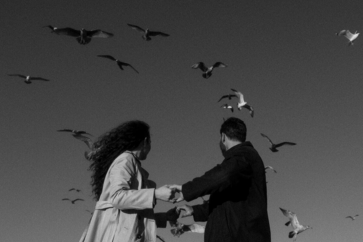 Couple holding hands while watching seagulls flying all around them in Newport, Rhode Island, by Andrea-Marie Photography