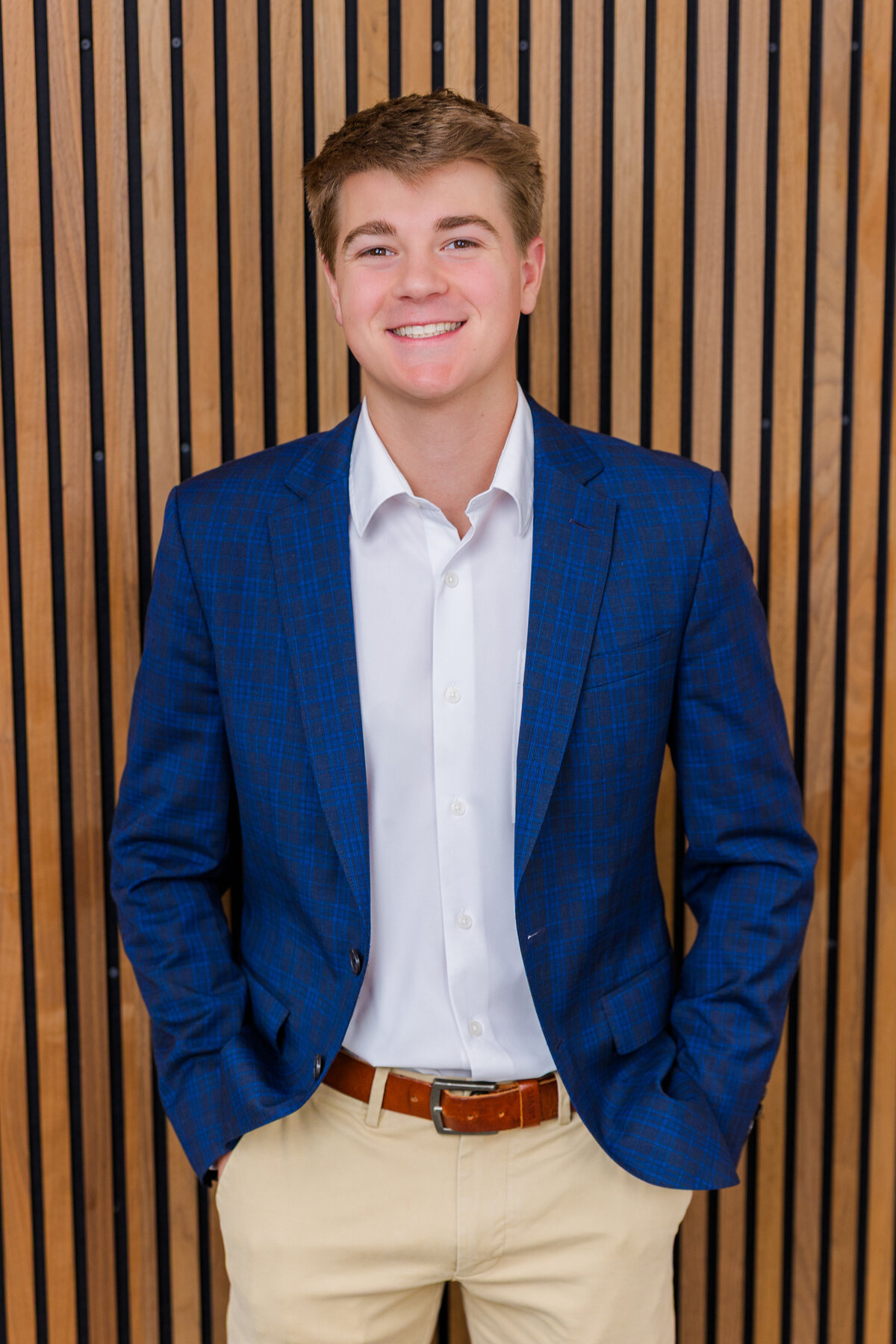 young professional portrait wearing a button up shirt, a blue jacket and light beige pants standing hands in pockets in front of a brown background in studio