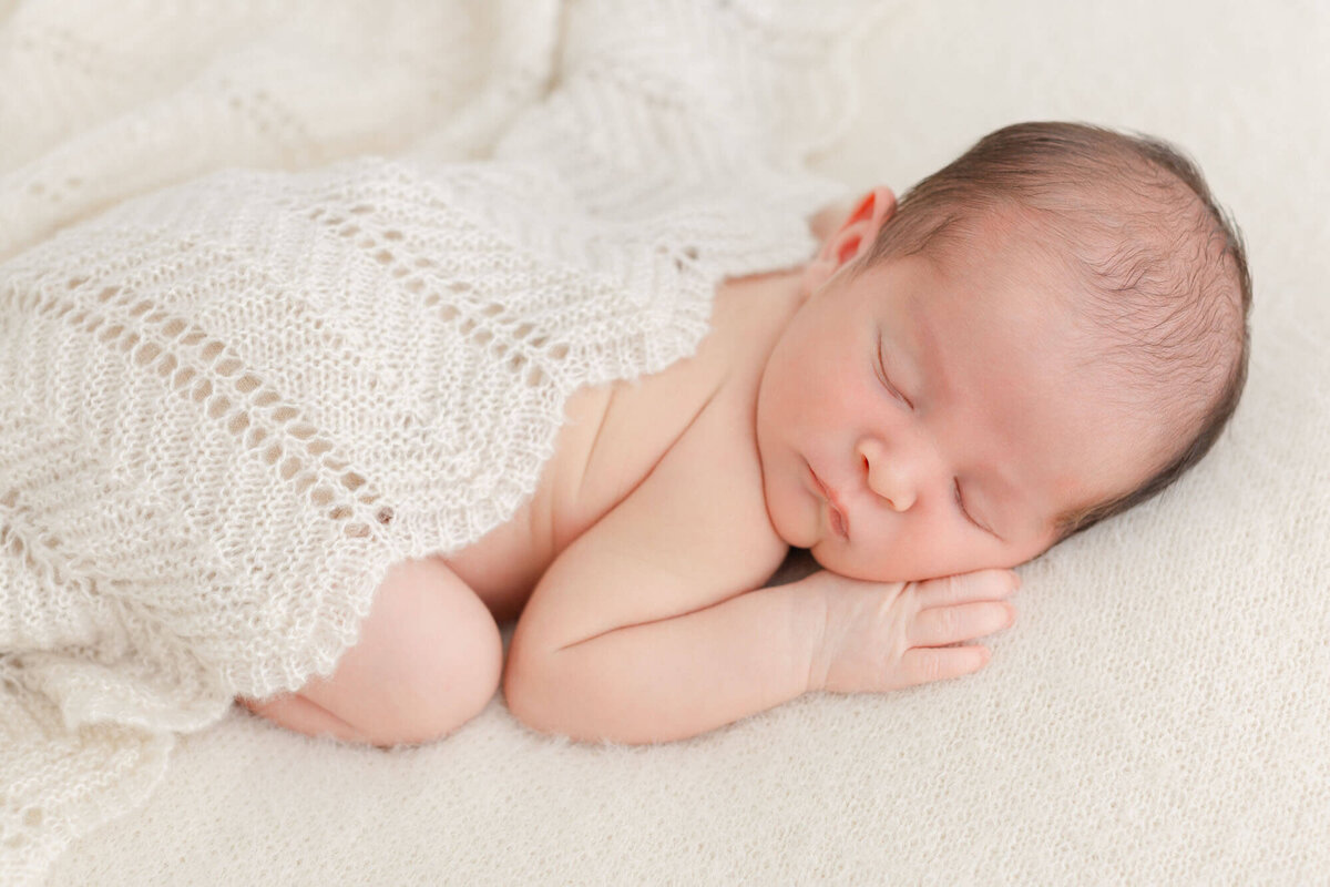 Newborn baby sleeping on belly on a beige backdrop with a crochet beige blanket draped over back. Hand is up by his face.