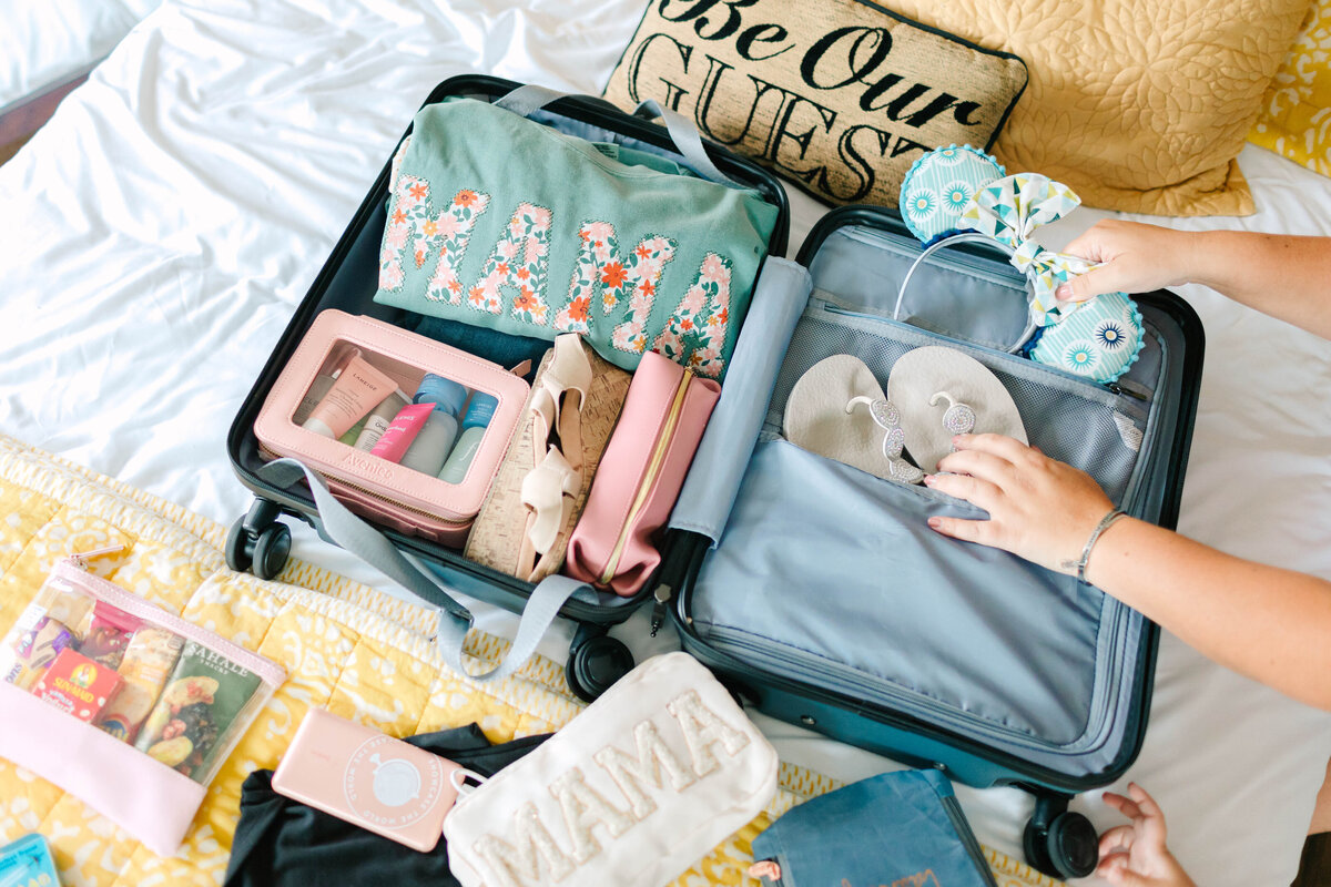 An open suitcase on a bed is neatly packed with travel essentials, including a "MAMA" sweatshirt, flip-flops, toiletry bags, and a pair of Mickey Mouse ears. A hand places the ears inside, while other items like a phone, snacks, and a travel pouch are spread out on the yellow quilted bedspread. A decorative "Be Our Guest" pillow rests against the headboard.