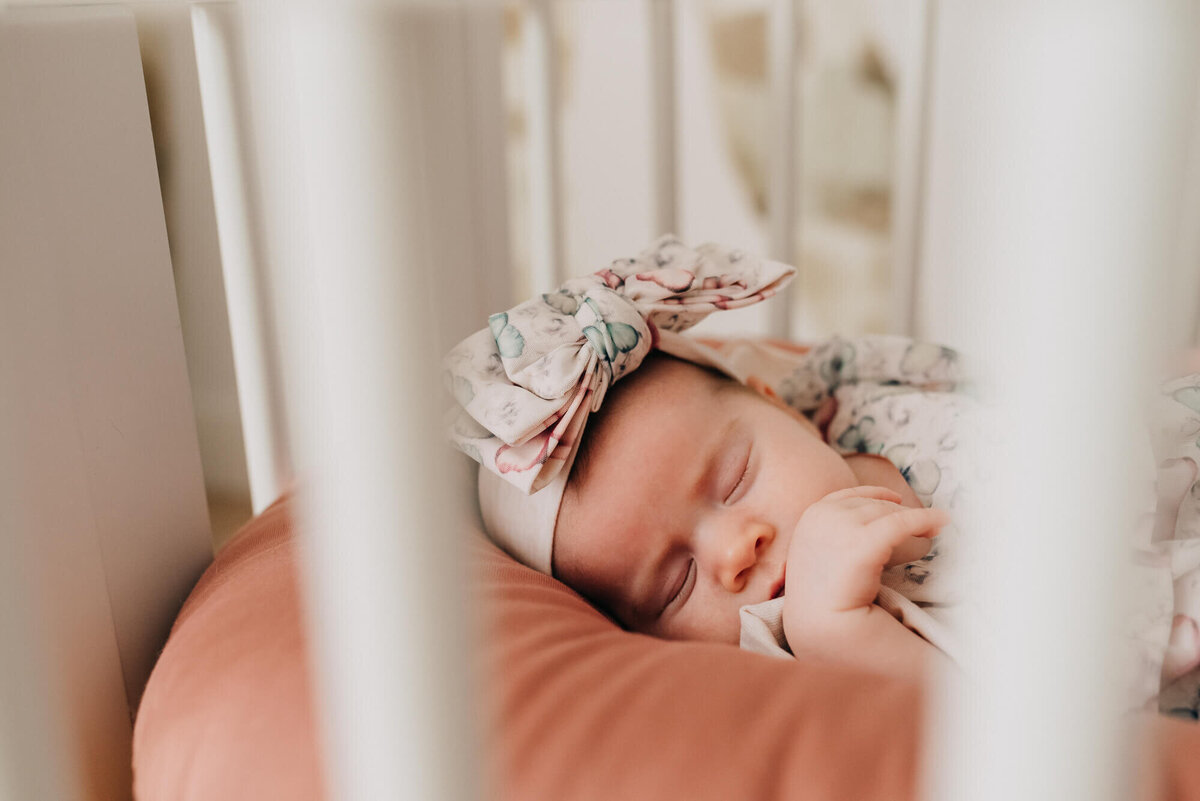 A closeup of a baby girl wearing a floral bow sleeping in a rose colored snuggleme in her crib