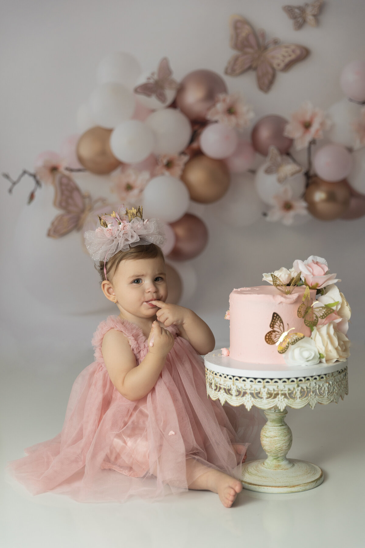 A toddler girl in a pink dress tastes the icing on her pink cake in a photo studio