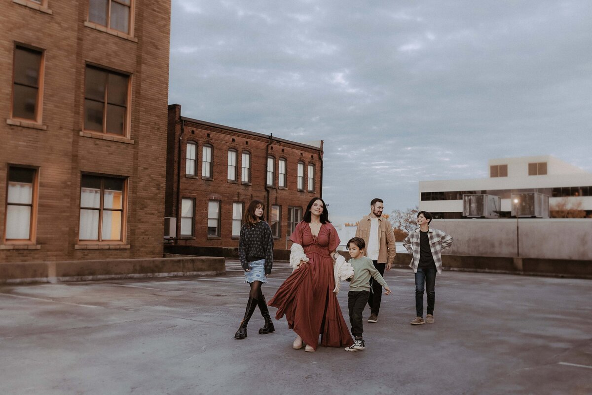 A group of people, including adults and children, walk together on a rooftop. They're casually dressed. Background includes brick buildings and a cloudy sky.