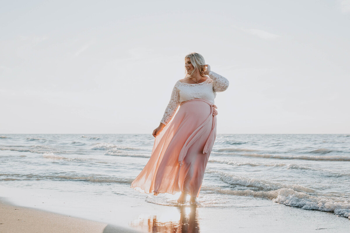 Expectant mom in Grand Bend beach maternity photoshoot. Mom is standing on the shoreline at golden hour in long pink dress and cream top. Her skirt is blowing in the wind and she is touching her hair.