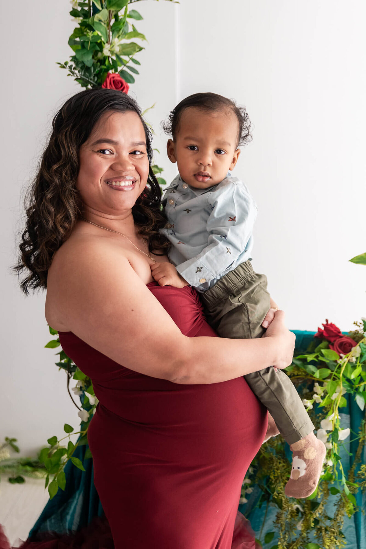 A pregnant woman in a red dress lovingly holds her young son, showcasing a beautiful moment of family connection.