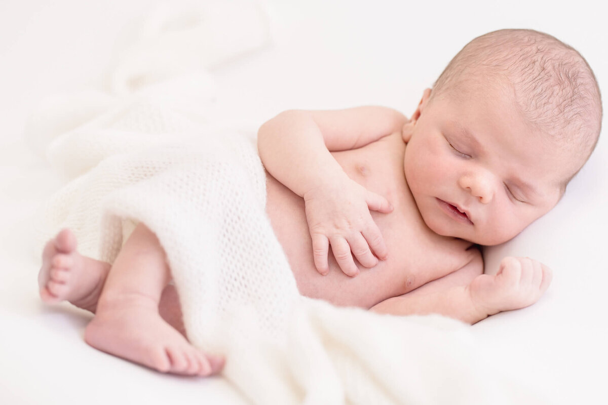 A newborn baby sleeps peacefully on a soft, white blanket. The baby is unclothed except for being partially covered by the blanket, with one hand gently resting on their chest. The background is a soft, light color.