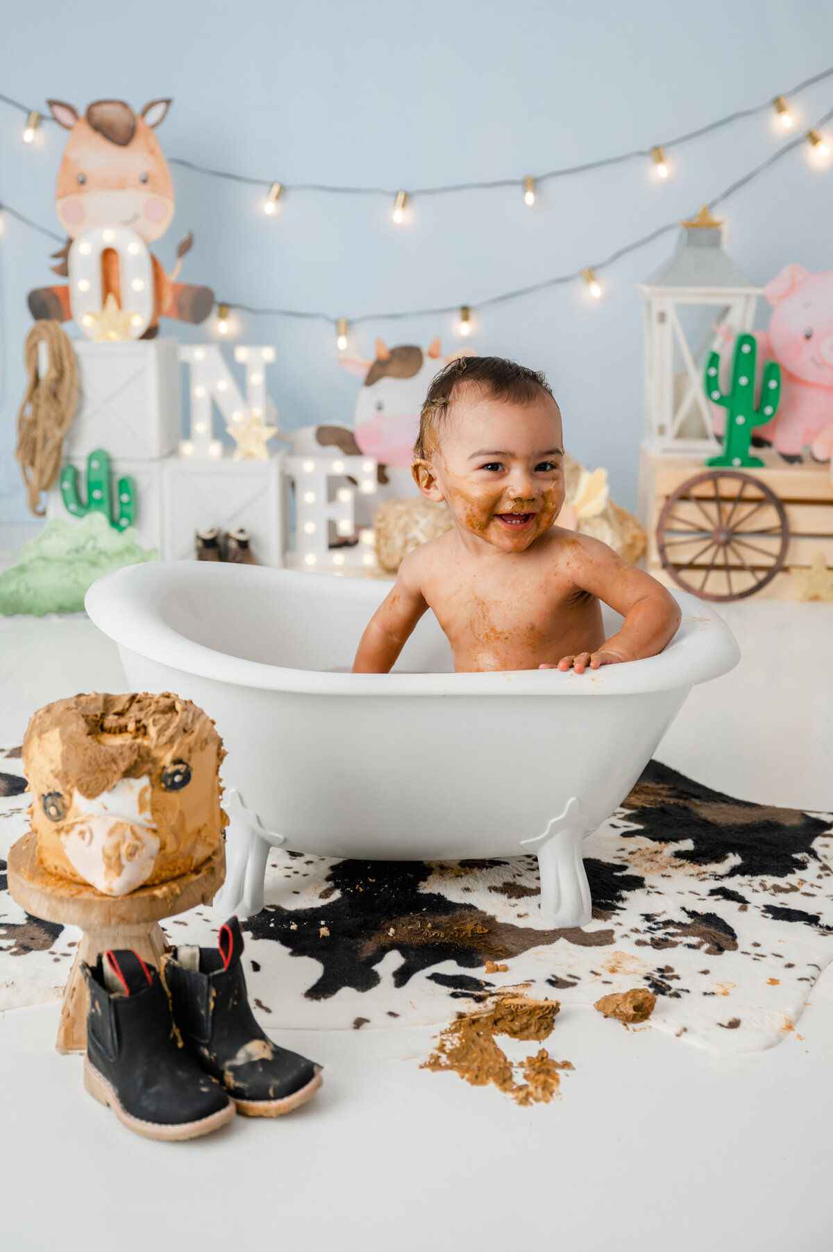 A smiling young boy sitting inside a mini tub, surrounded by a cowboy-themed backdrop with pieces of cake everywhere. 