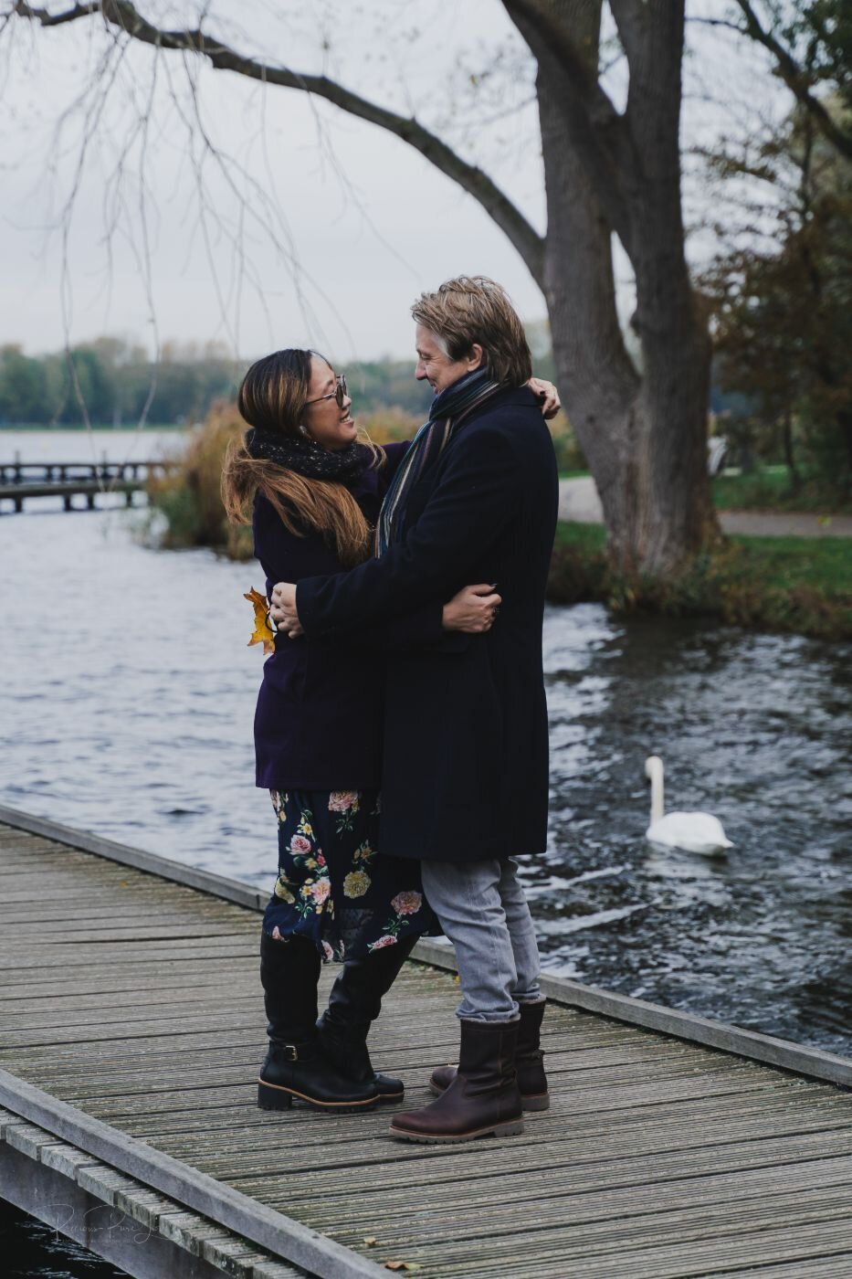 A romantic, full-body portrait of a couple embracing by the water on a cloudy day. The woman, with long brown hair, is wearing glasses, a purple coat, and a floral skirt. She has her arms wrapped around the man, who is wearing a dark blue coat and a striped scarf. They are looking into each other's eyes, and a few ducks are visible on the water behind them.