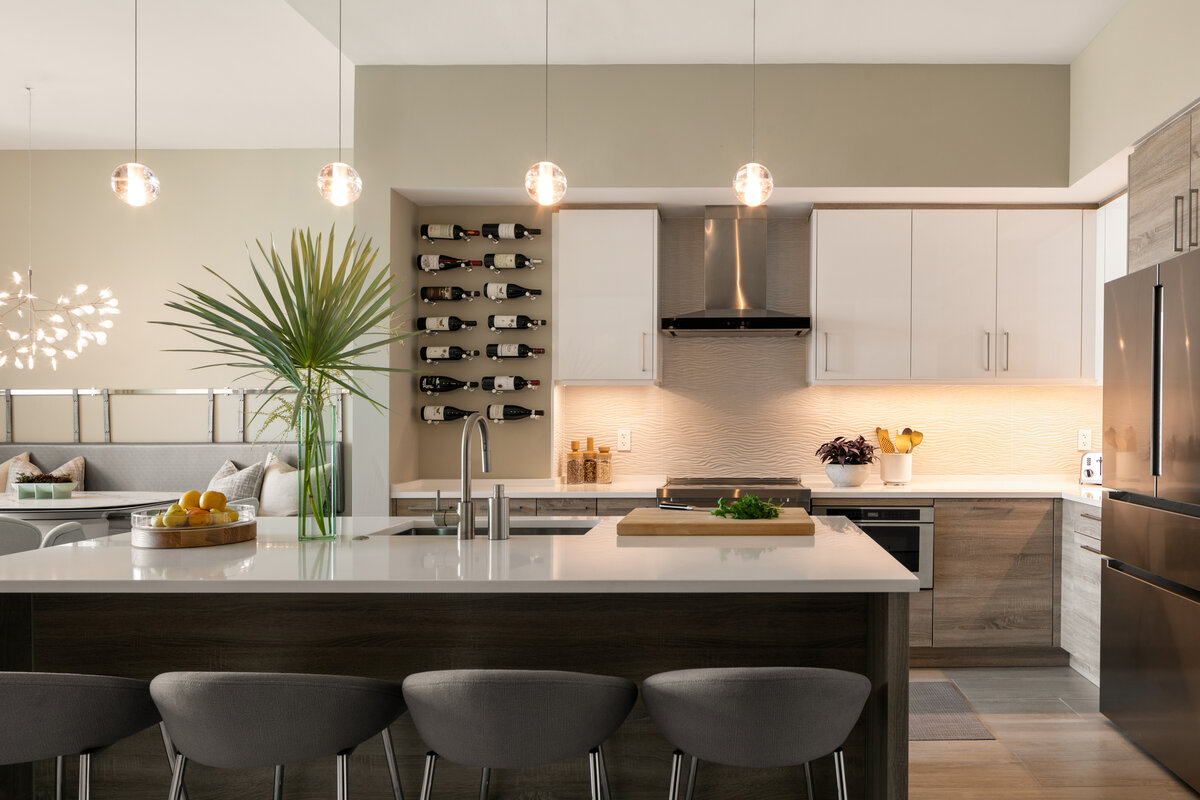 Feature photo for the Minky at the Mark gallery. Open concept kitchen living room, photo focus is the kitchen with silver appliances, white counters and grey wood cabinets and counters, white textured backsplash, and statement floating wine rack.
