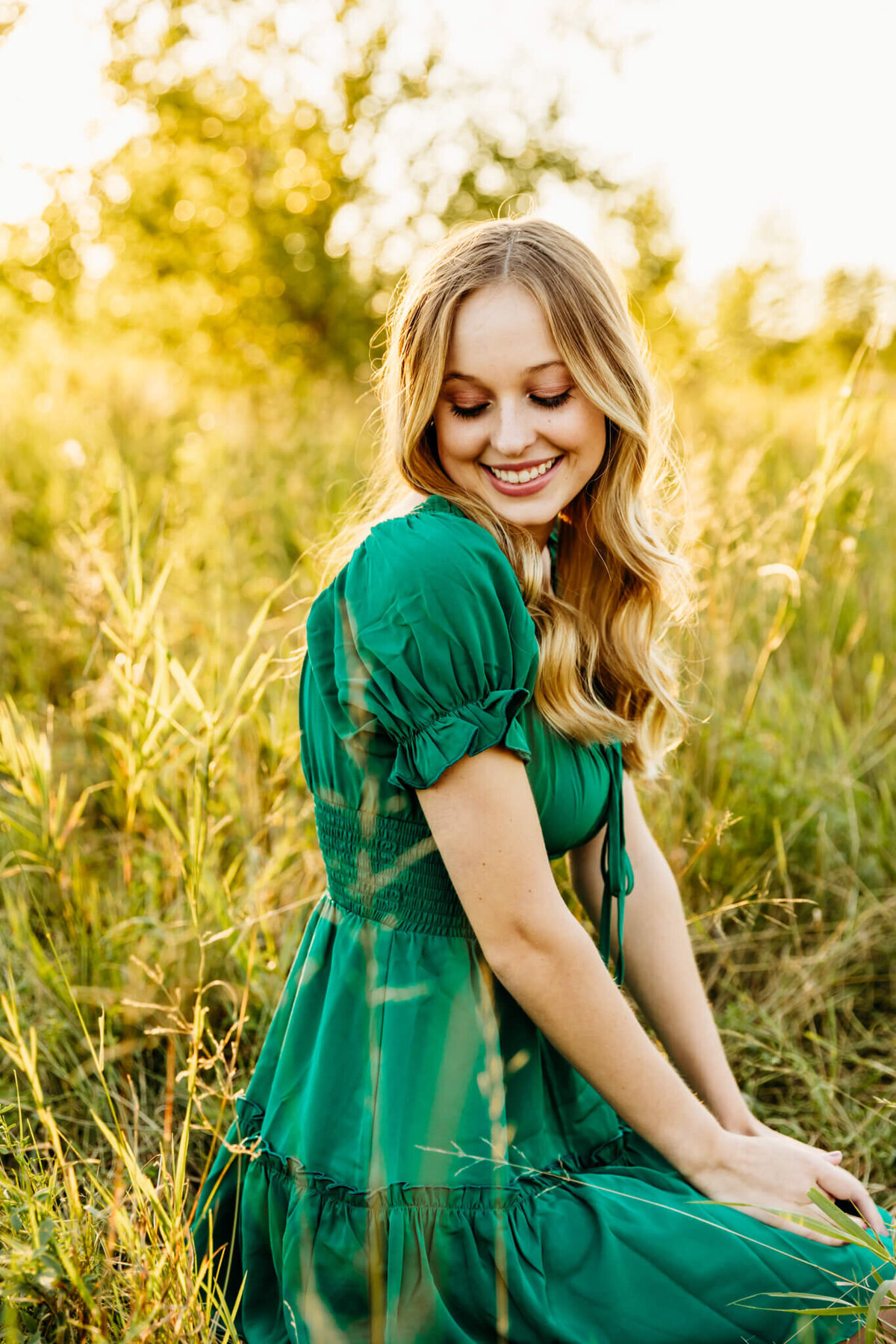 teenage girl in a green dress laughing as she glances down while sitting in a field near Oshkosh