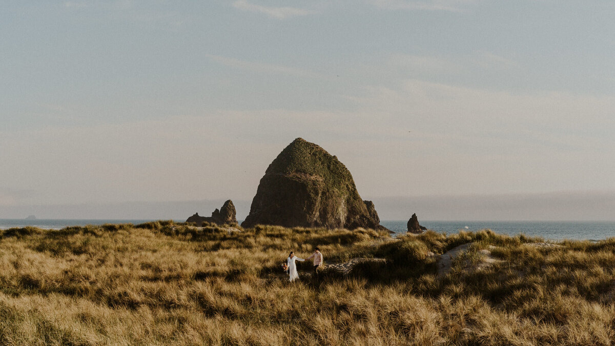 A couples walks through the sea grass infront of haystack rock. The