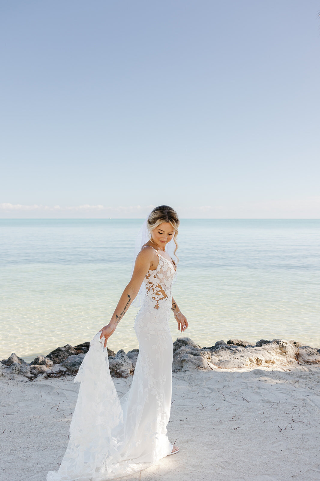 Ashley smiles while holding the train of her lace wedding dress on the sandy shore.