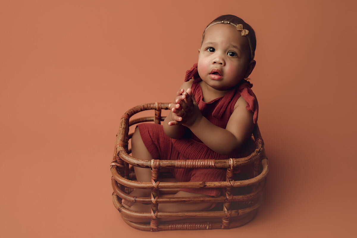 a one year old girl being shy in basket on a tan backdrop