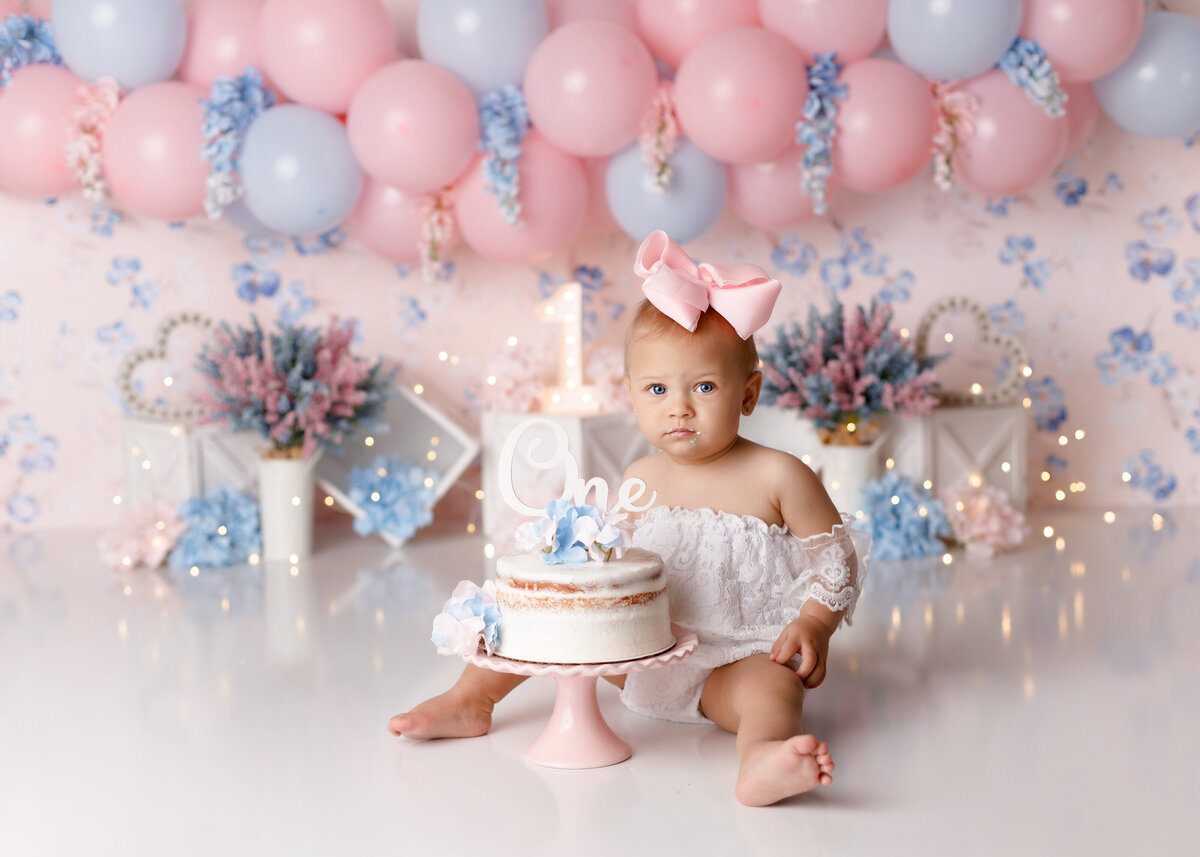 Pink and blue floral themed cake smash in West Palm Beach and Palm Beach Gardens Florida newborn and cake smash photography studio. Baby girl is wearing a lace off the shoulder white romper sitting behind a white decorated cake. In the background is a pale pink with light blue flowers surrounded by pink and blue florals with a soft pink and soft blue balloon garland.