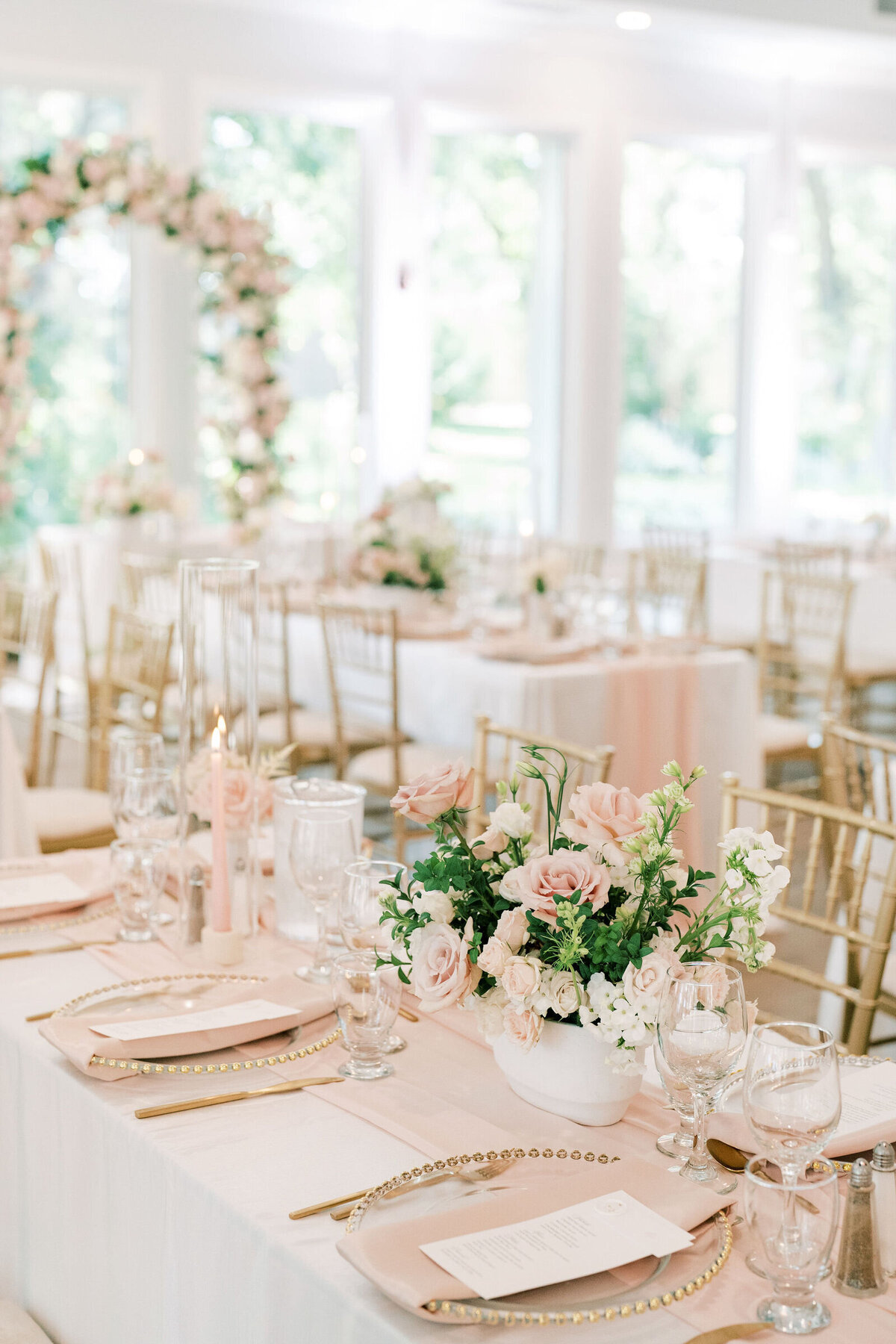 A wedding reception table setup with pink and white floral arrangements, candles, and elegant tableware is arranged in a bright room with gold chairs and a floral arch in the background, showcasing the exquisite touch of a full wedding planner.