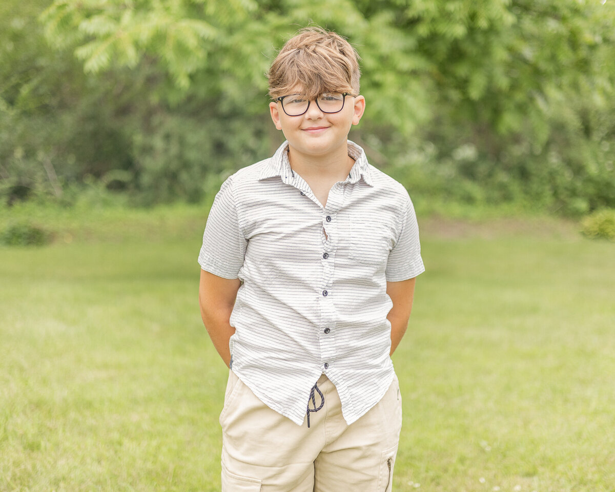 Younger son standing in the grass with a tree line behind him and his hands behind his back, looking at the camera smiling, wearing a light tan short sleeve button up shirt, khaki shorts, wearing glasses, and has short light brown hair
