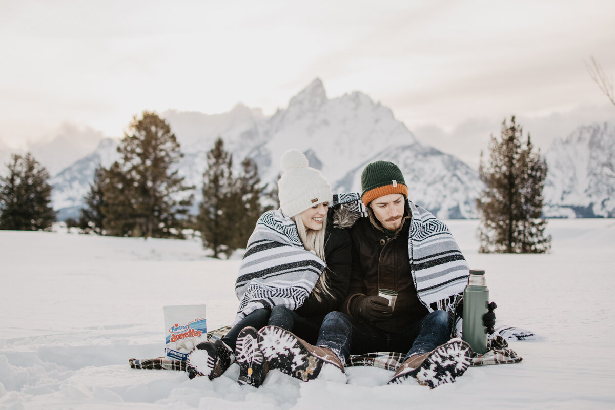 engaged couple sitting on a blanket in the snow at Jackson Hole while they also have a blanket around their shoulder as they cuddle up for their winter engagement session