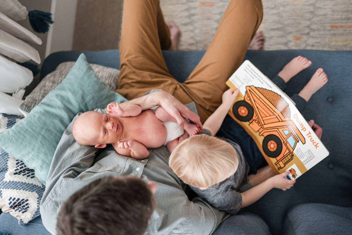 A father sitting on a couch with his newborn baby in his arms, while the toddler sits beside him, both looking at a book about a dump truck.