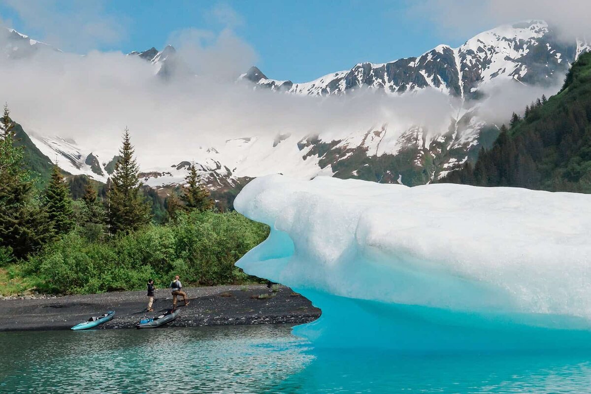 A man and woman and their guide are visible on a black pebble beach during day two of their luxury elopement in Alaska.  On the edge of the water are kayaks, and slightly out of focus on the right edge of the frame is a huge blue-tinted white icebergs floating in Bear Lake. Behind the beach is a row of green grasses and pine trees, behind that tall, craggy snow-covered mountains fill the frame 