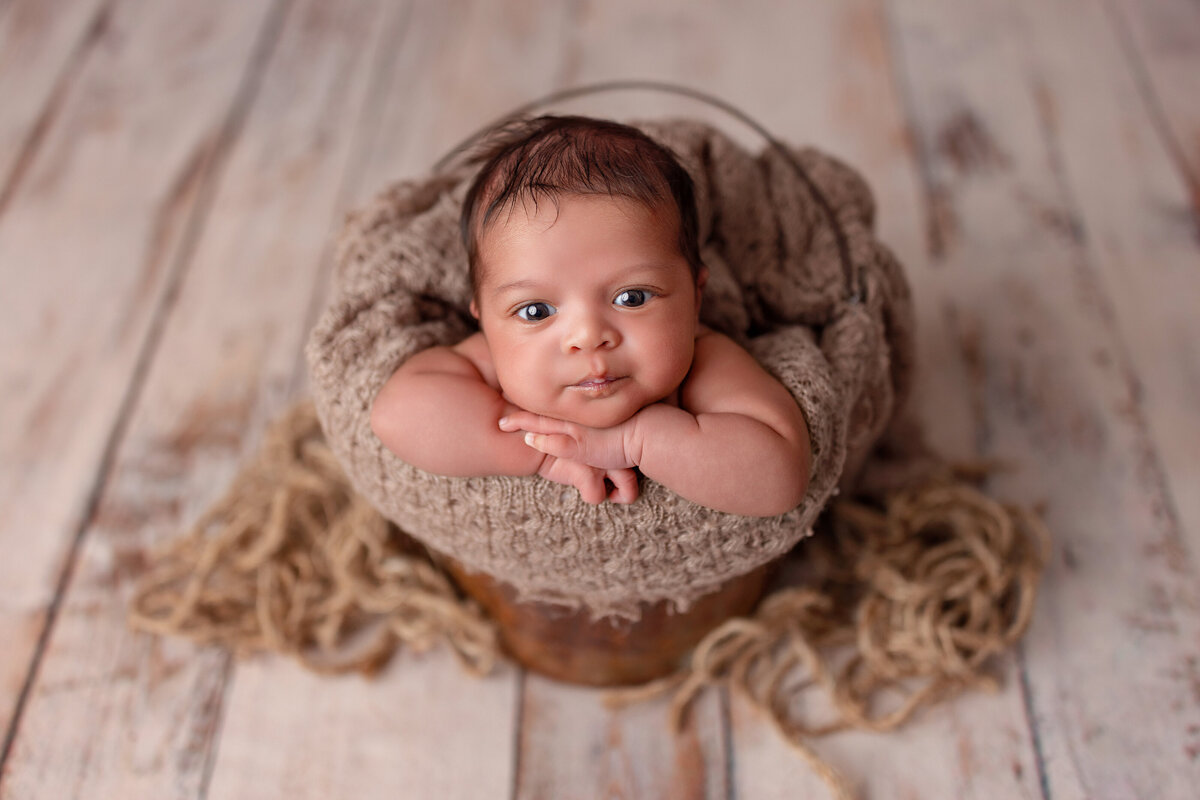 Newborn baby resting in a rustic wooden bucket on a wood floor with cozy textured blankets.