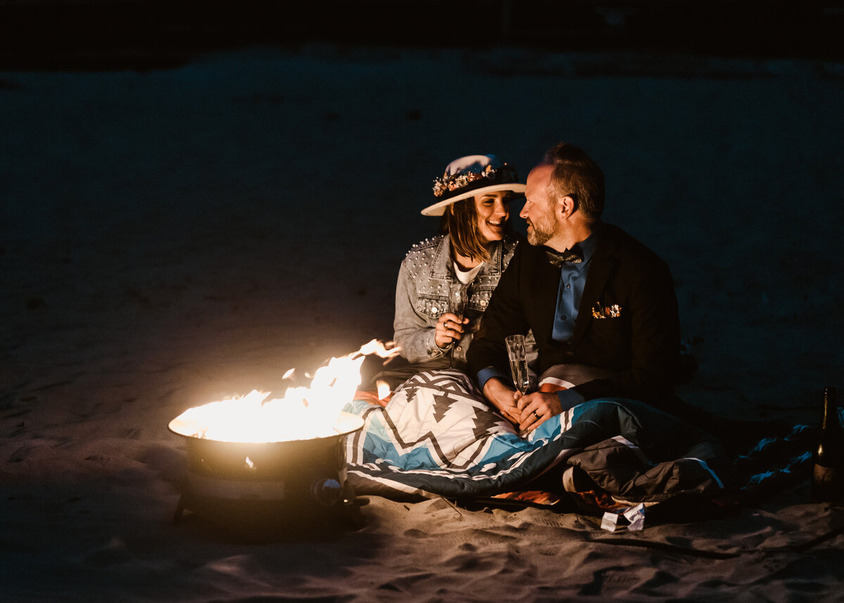 At the end of their Oregon coast elopement, a bride and groom snuggle up by a fire. They toast champagne as they cuddle under their camping blanket.