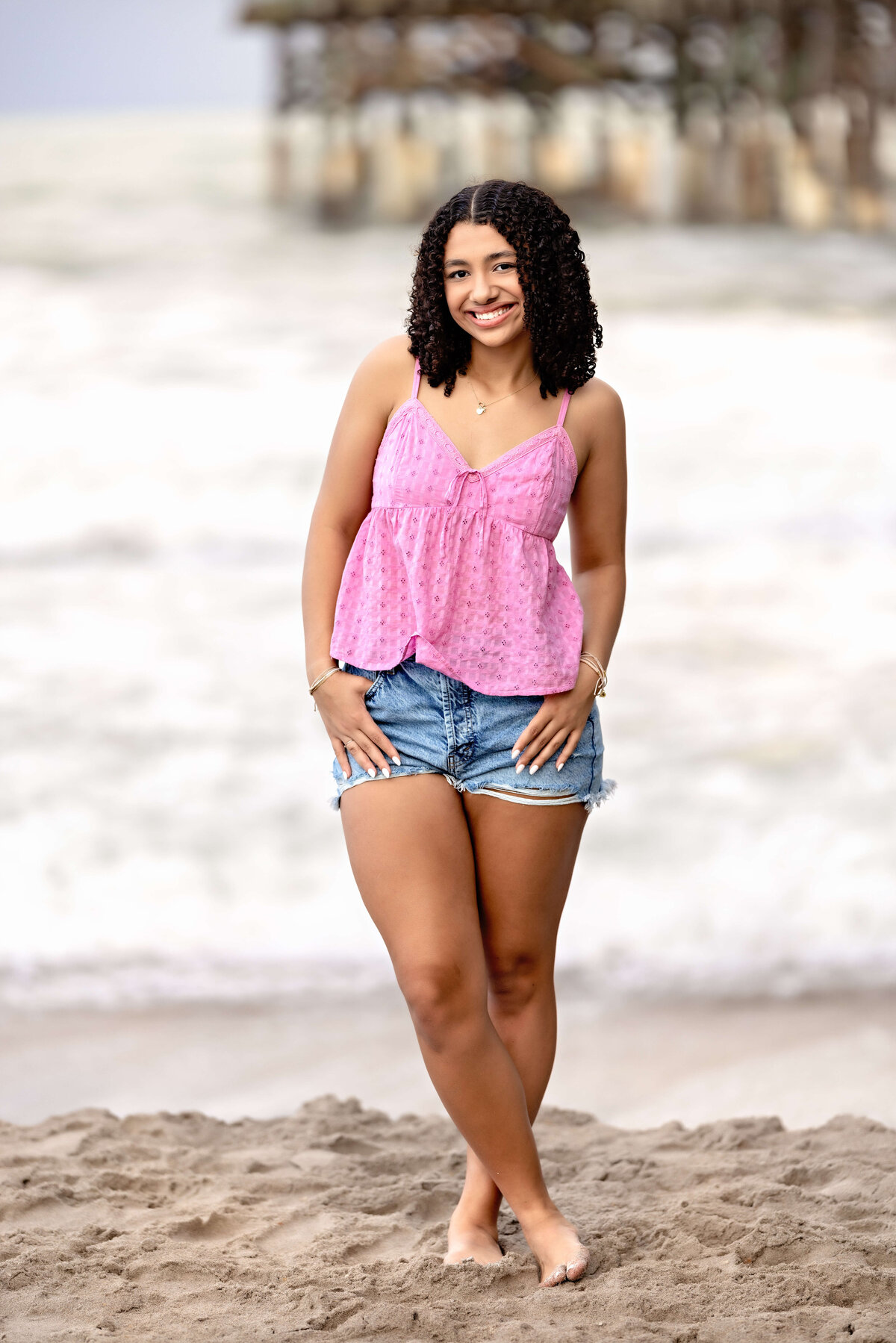 Senior girl smiling with hands in pockets, standing at the shoreline with waves and storm clouds creating a dreamy Cocoa Beach backdrop.