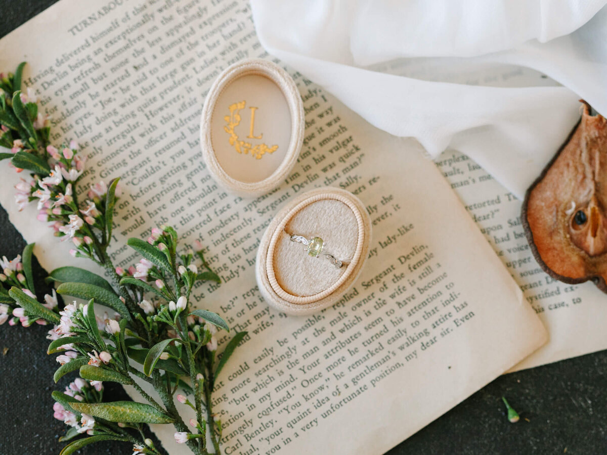 An ring with a green gemstone sits in an open velvet box on vintage book pages. Nearby are sprigs of small delicate flowers.