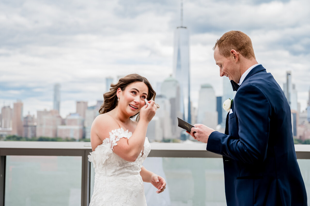 A bride wiping her tears as her groom reads his vows to her while they stand on a rooftop overlooking the NYC skyline