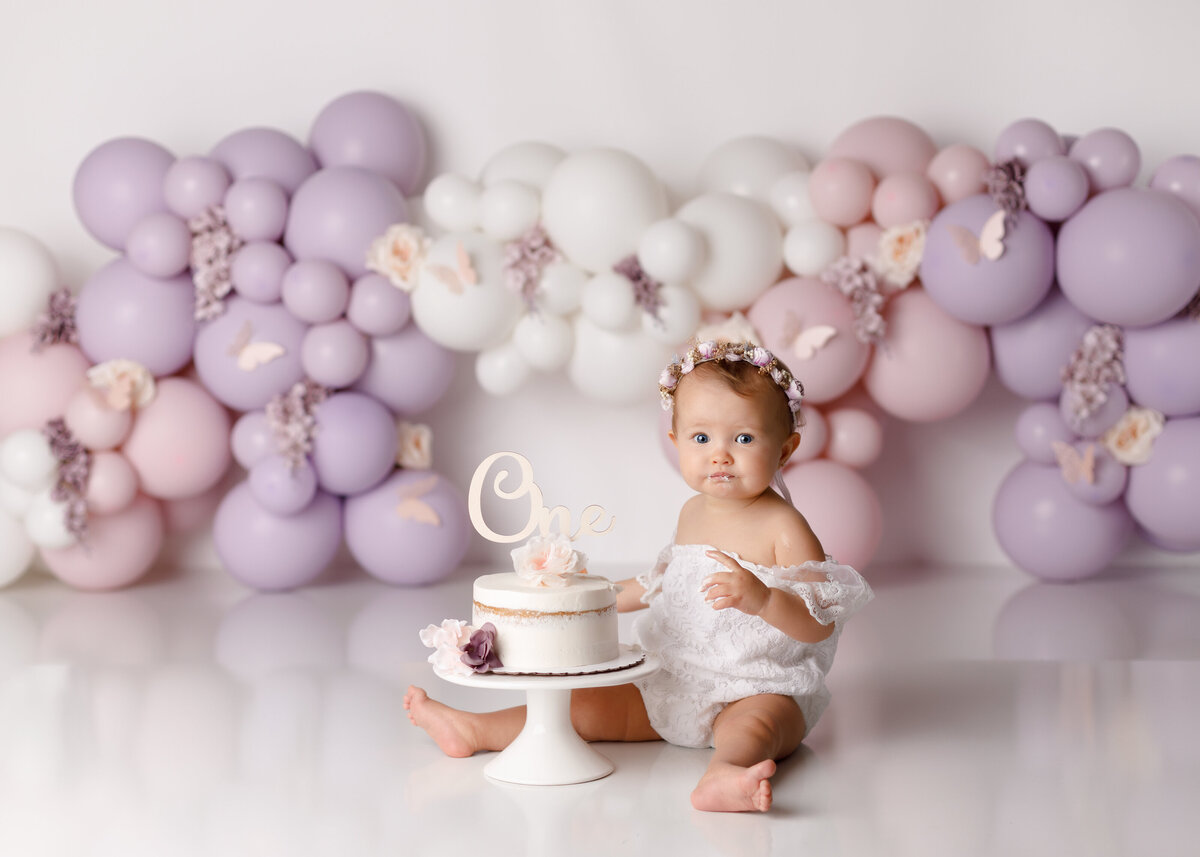 Soft pink and lavender balloon garland cake smash in West Palm Beach Florida portrait studio. Baby girl is wearing an off the shoulder white lace romper with a floral crown sitting beside a white naked cake decorated with flowers. The background is a custom-made balloon garland and lavender soft pink and cream with florals scattered around.