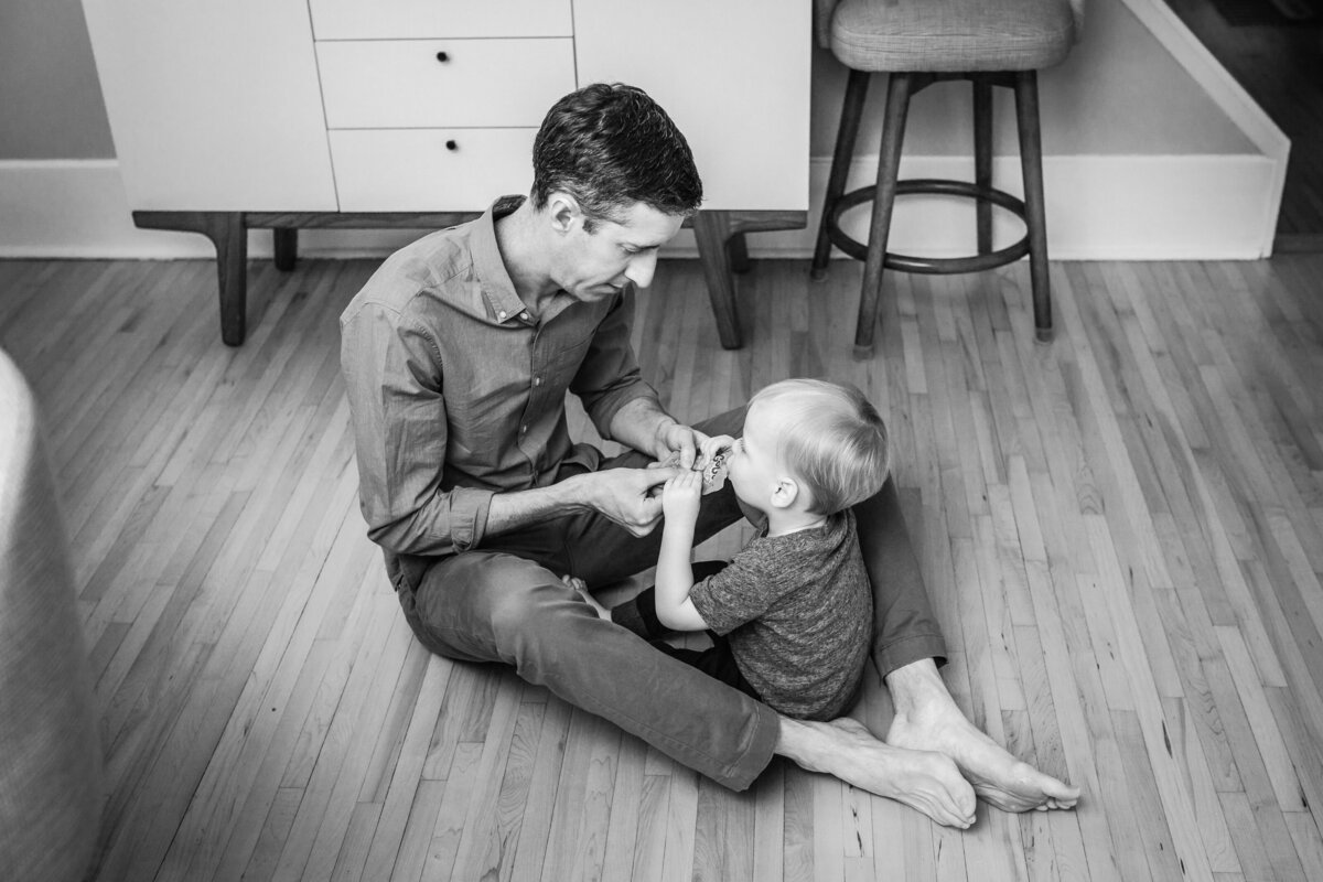 A father sitting on the floor with his toddler, helping him with a small activity or toy in a cozy, light-filled room.