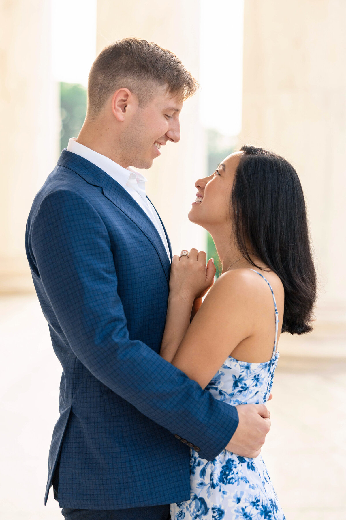 A couple stands in an elegant embrace, smiling at each other. The man wears a blue suit, and the woman is in a blue and white floral dress. They stand against a light background with columns.