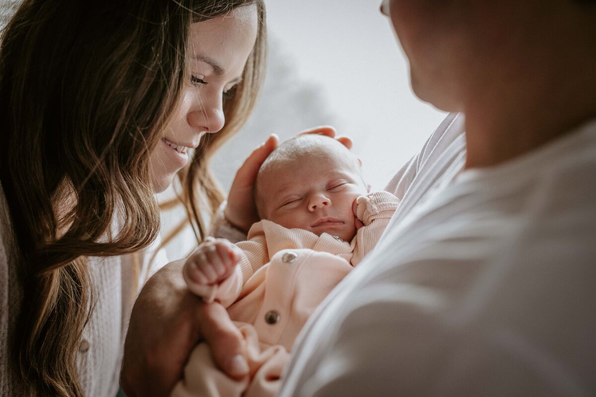 In home newborn session - Dad holding baby and mom facing him and touching baby's head. Image is shot over dad's shoulder - dad is blurred and mom and baby are focus of the image.
