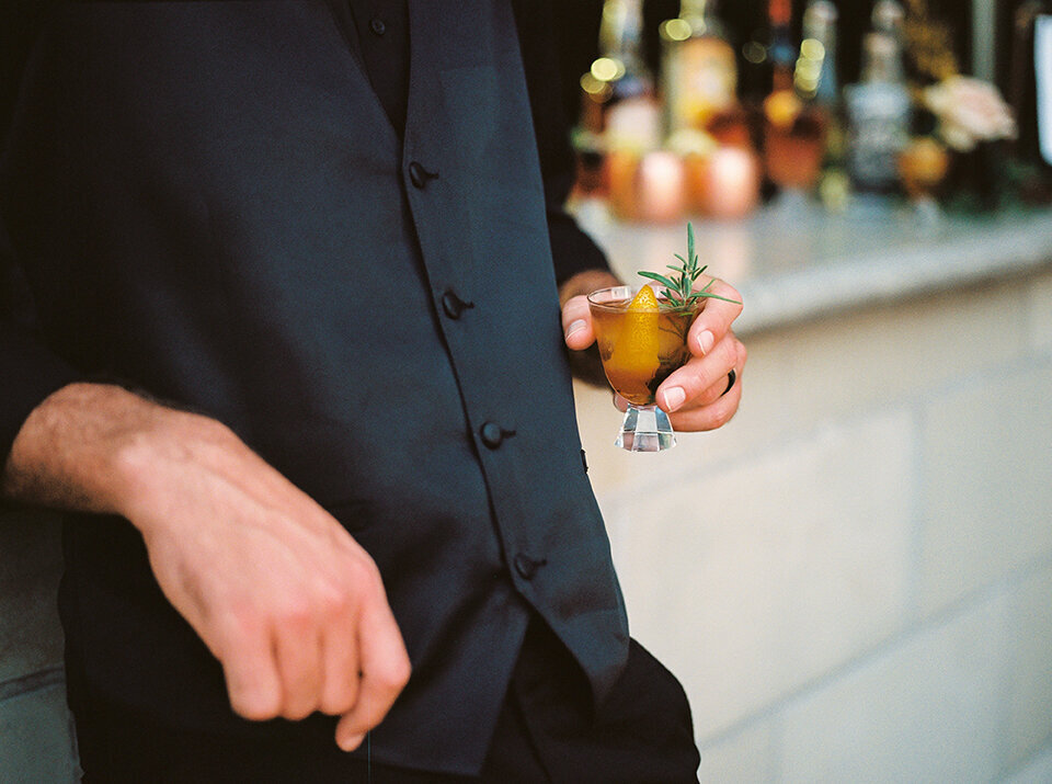 The groom wearing a black suit holds a cocktail drink in hand while leaning against the bar.