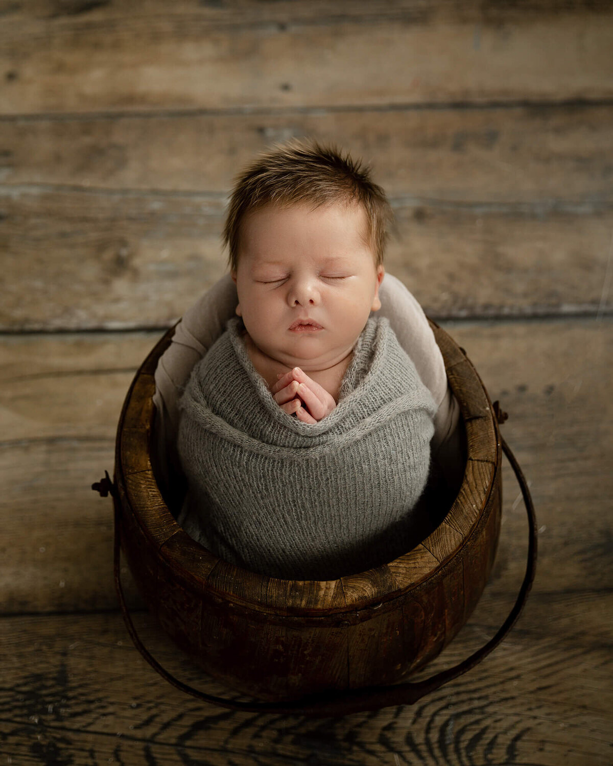 Newborn boy with lots of light brown hair sleeping in a wood bucket with his hands together like he's praying.