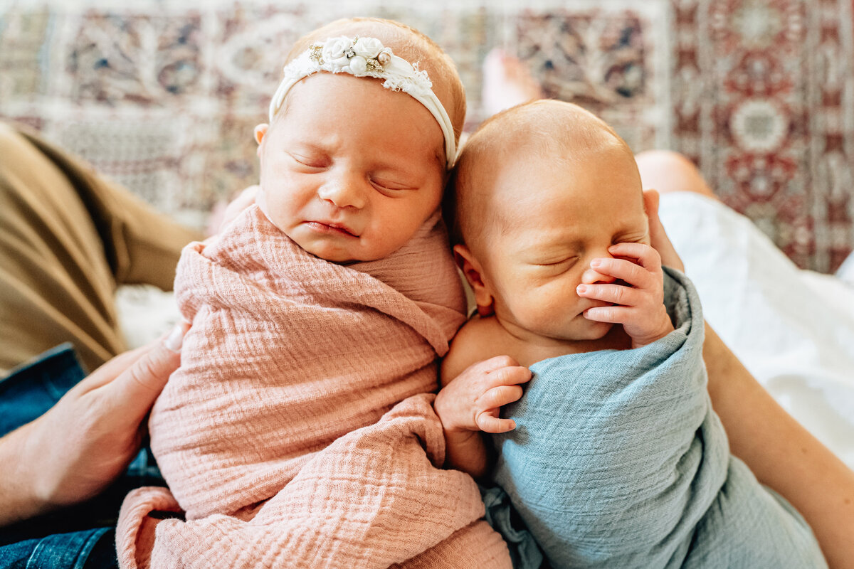 A close-up color image of two newborn twins, one wrapped in a pink swaddle with a floral headband, and the other in a blue swaddle. The babies are sleeping peacefully, nestled closely together in their parents' arms.