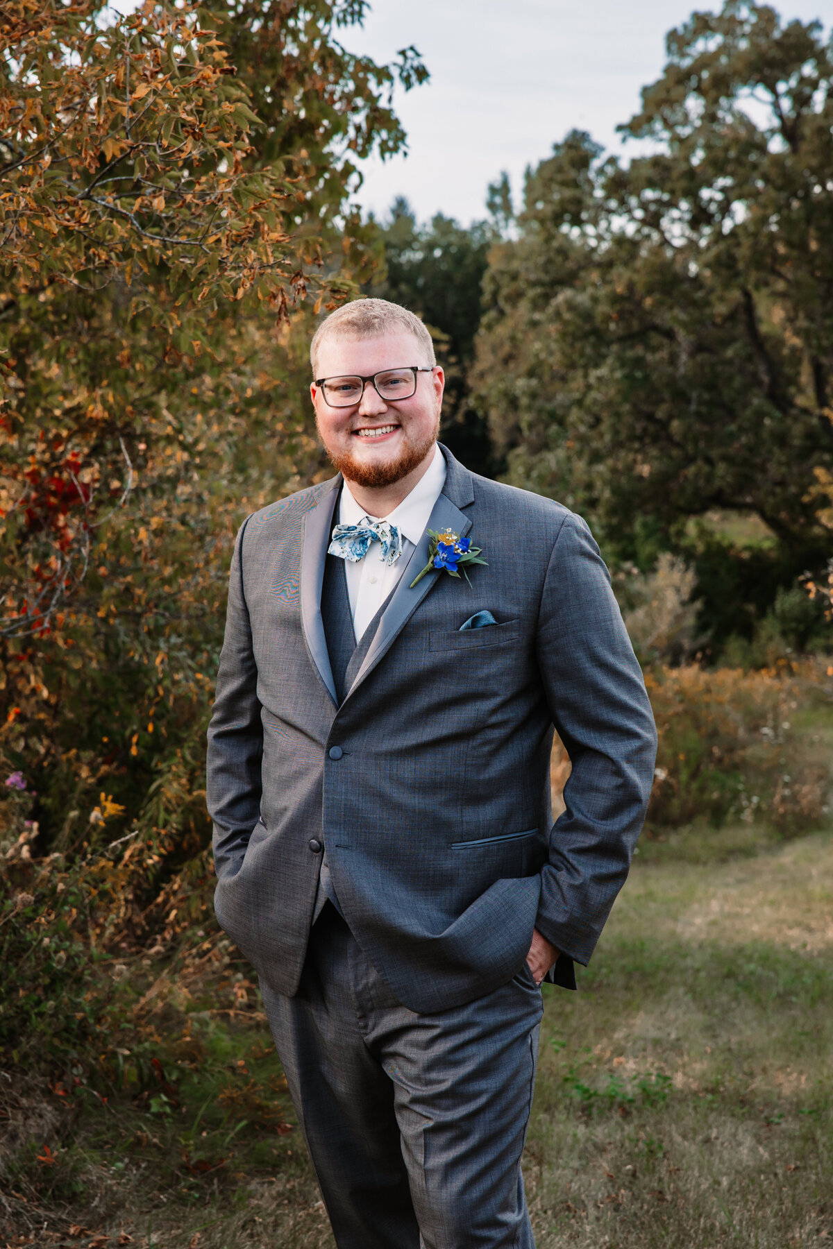A groom in a grey suit and blue bowtie stands in a trail with hands in pockets