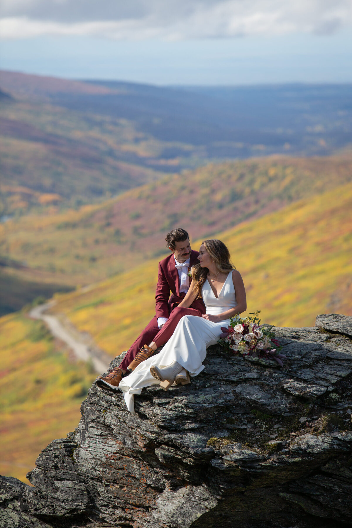 A bride and groom sit together on a rock with a flower bouquet resting next to the bride's leg.