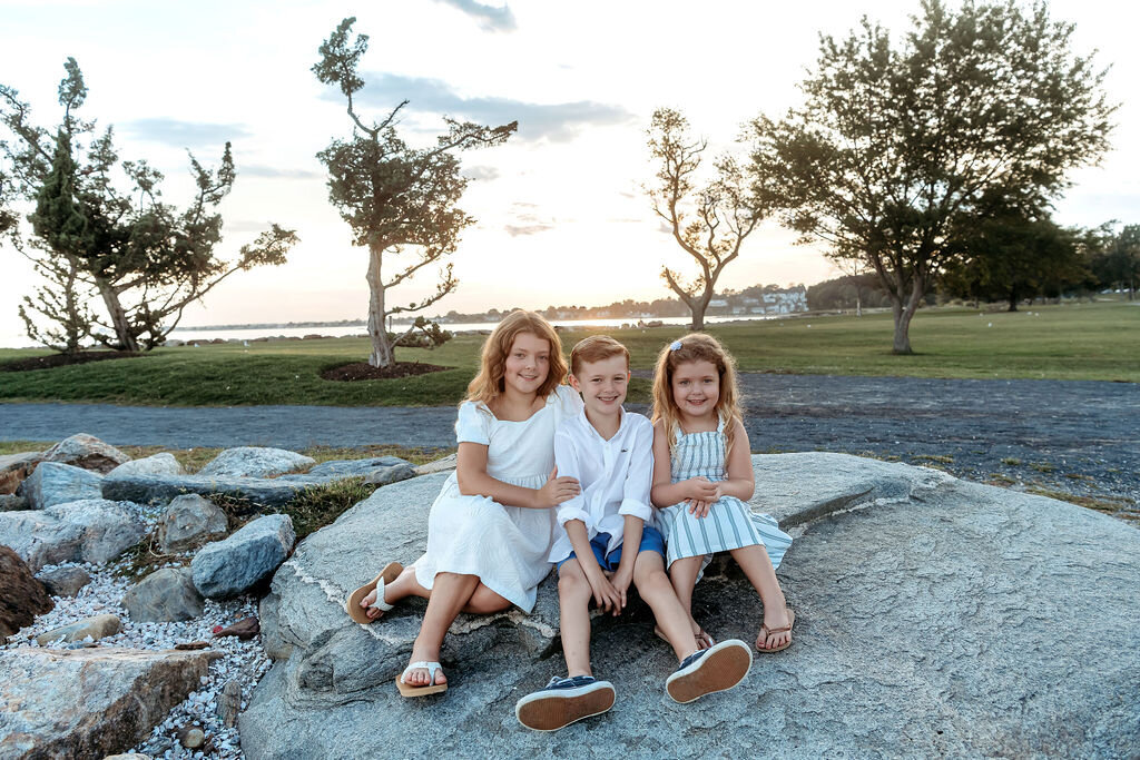 Three siblings in coordinating blue and white outfits posing on rocks during Connecticut family photographer waterfront session