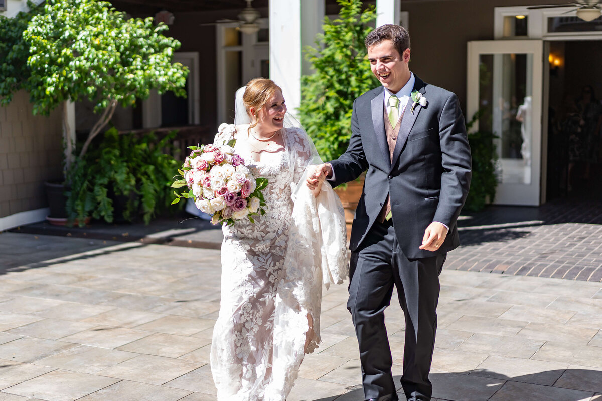 A smiling bride in a white lace gown holding a bouquet walks hand-in-hand with a groom in a black suit and gold tie outdoors on a sunny day.