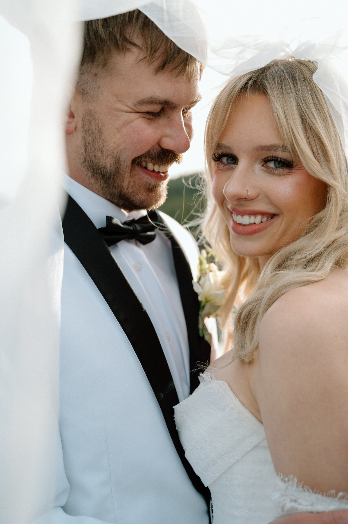 Groom smiles at camera while bride looks over shoulder with veil flowing in Banff.